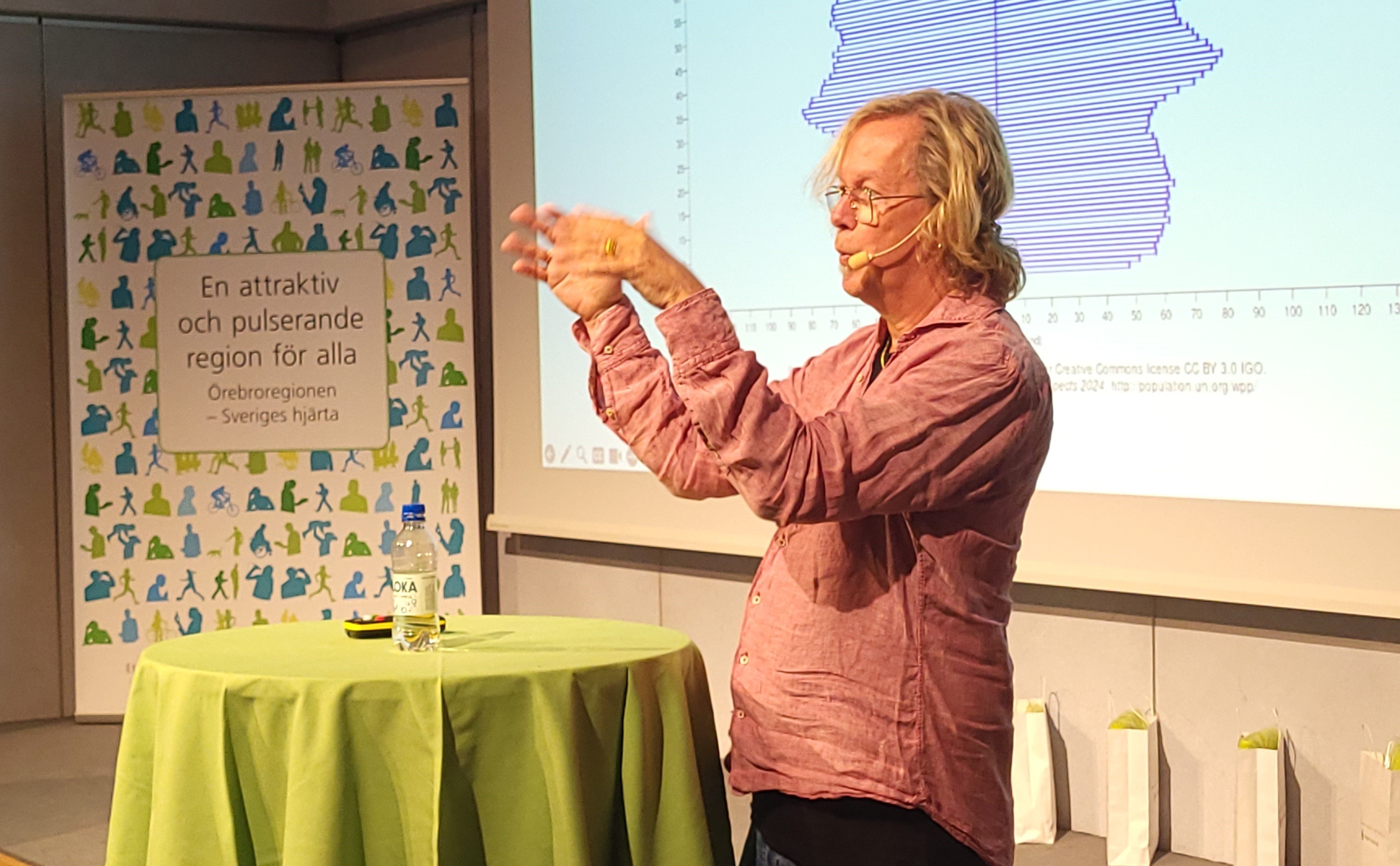 En man med halvlångt, blont hår i rosa skjorta gestikulerar. Bakom honom en presentation som visar en befolkningspyramid.
