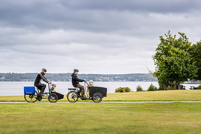 Två personer på lastcykel där det sitter barn i lådan. De cyklar på en cykelväg över en gräsmatta, invid en sjö.