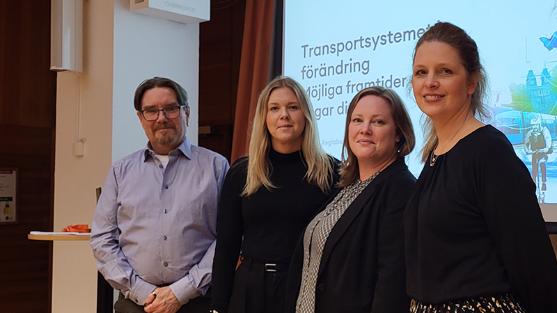 Fyra personer framför en stor digital skärm. De tittar in i kameran.