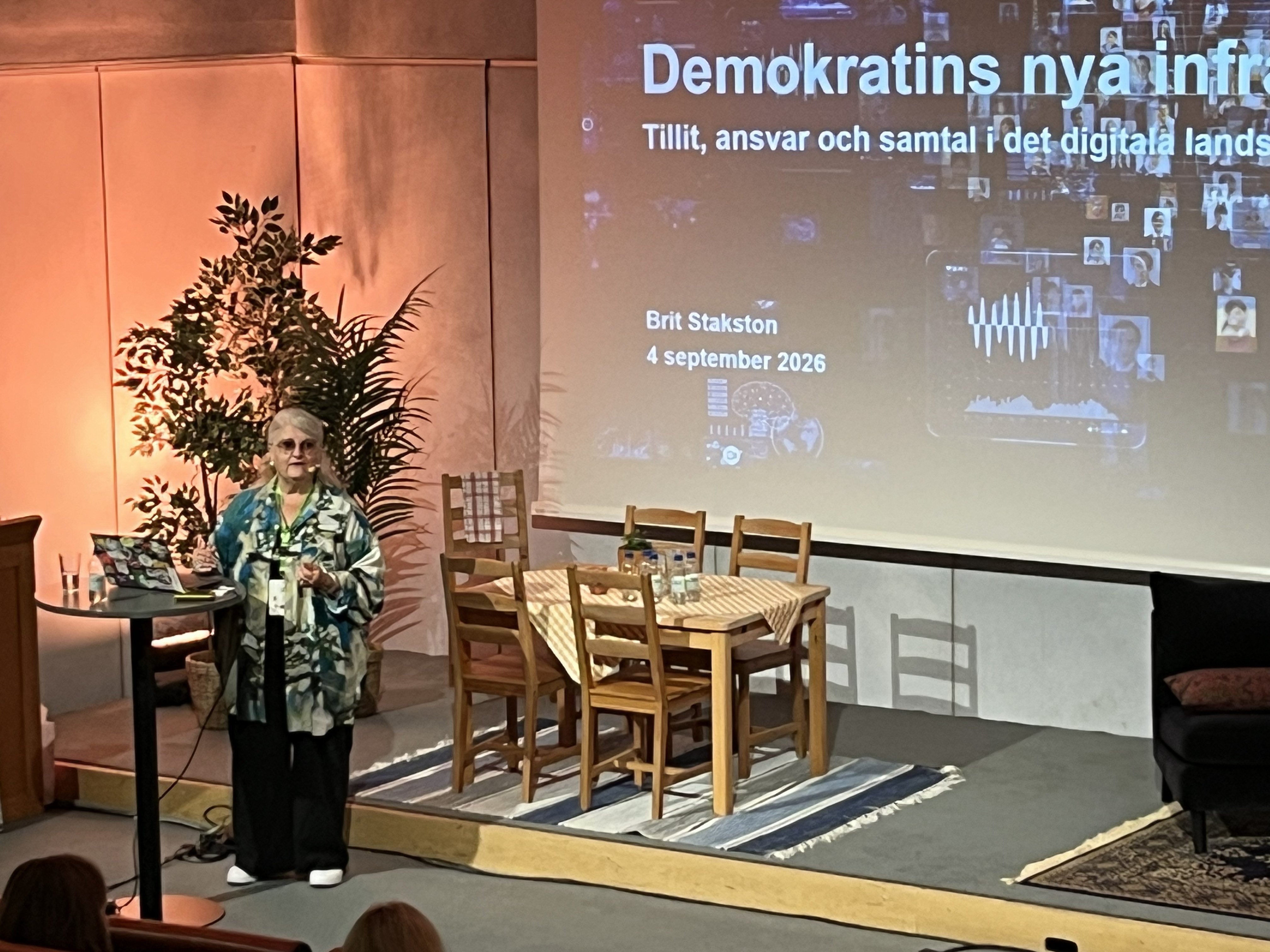 En kvinna föreläser. På scenen ett köksbord med stolar. På skärmen står Demokratins nya infrastruktur.