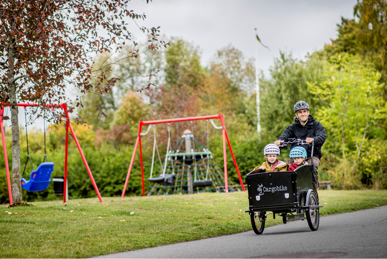 En man med två barn i lådcykel cyklar förbi en lekpark.