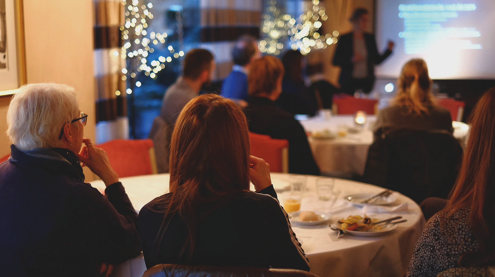 Flera personer sitter runt dukade frukostbord och lyssnar på föreläsare
