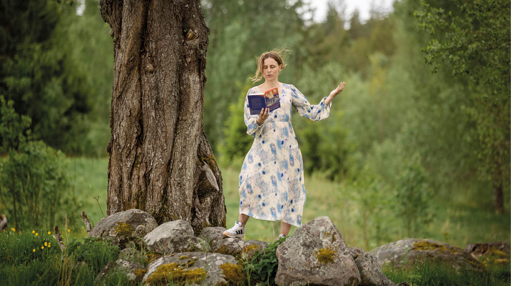 Kvinna i ljus långklänning läser högt ur en bok stående vid en tjock trädstam på en stenmur, djup skog i bakgrunden