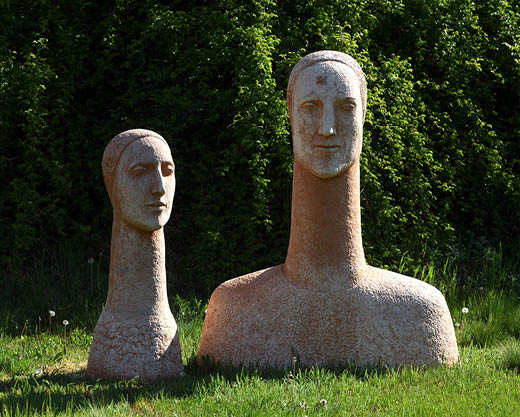 Två torso-skulpturer med höga halsar i lera står på en gräsmatta