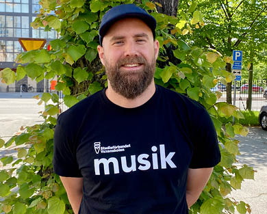 Man med skägg och keps, det står musik med stora bokstäver på hans svarta t-shirt