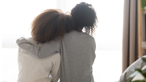 kram - en kvinna lägger armen om en annan . De sitter på en säng och har ryggen mot oss.