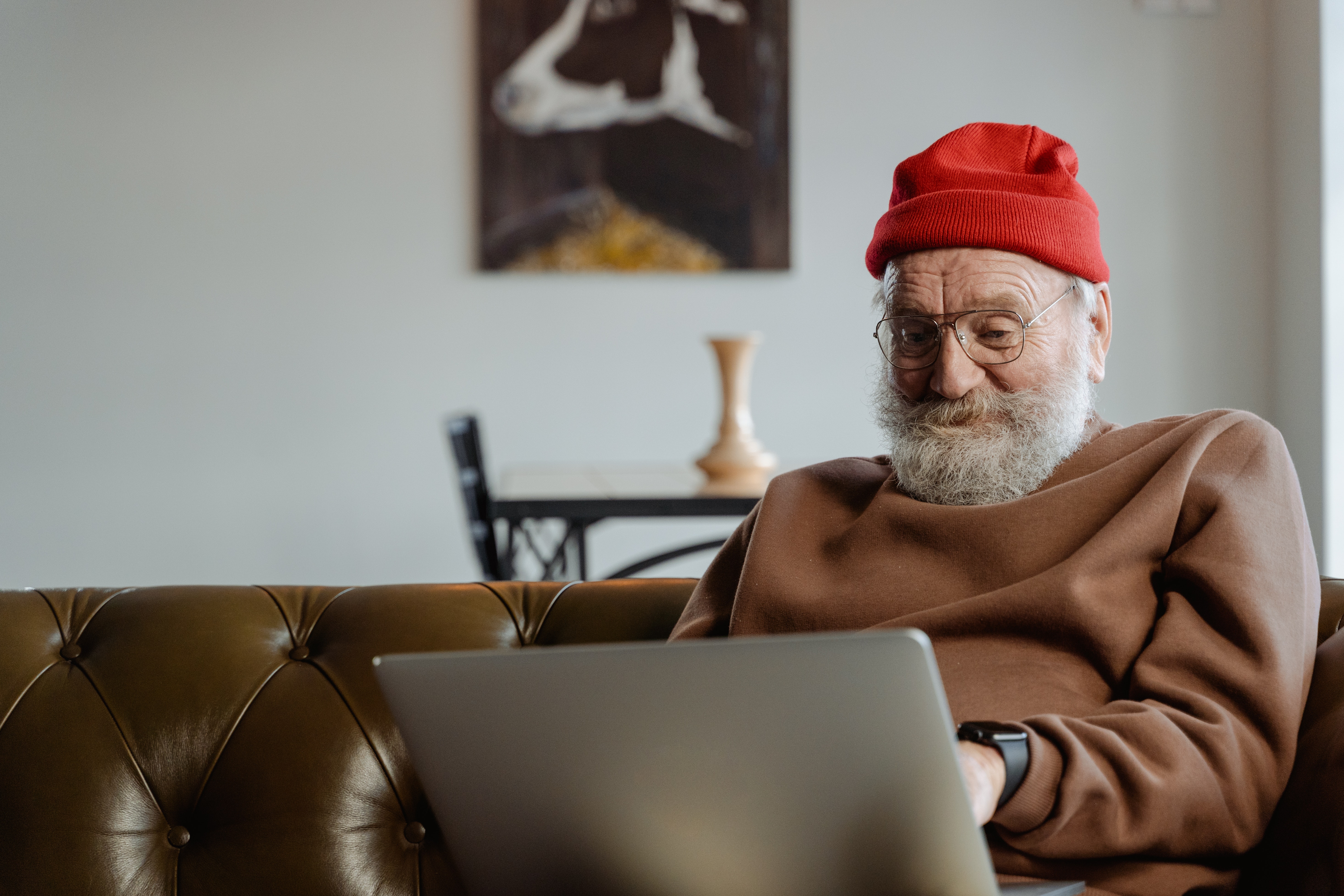 En äldre herre med grått hår och röd mössa sitter i soffan med en laptopdator i knät. Han ler finurligt.