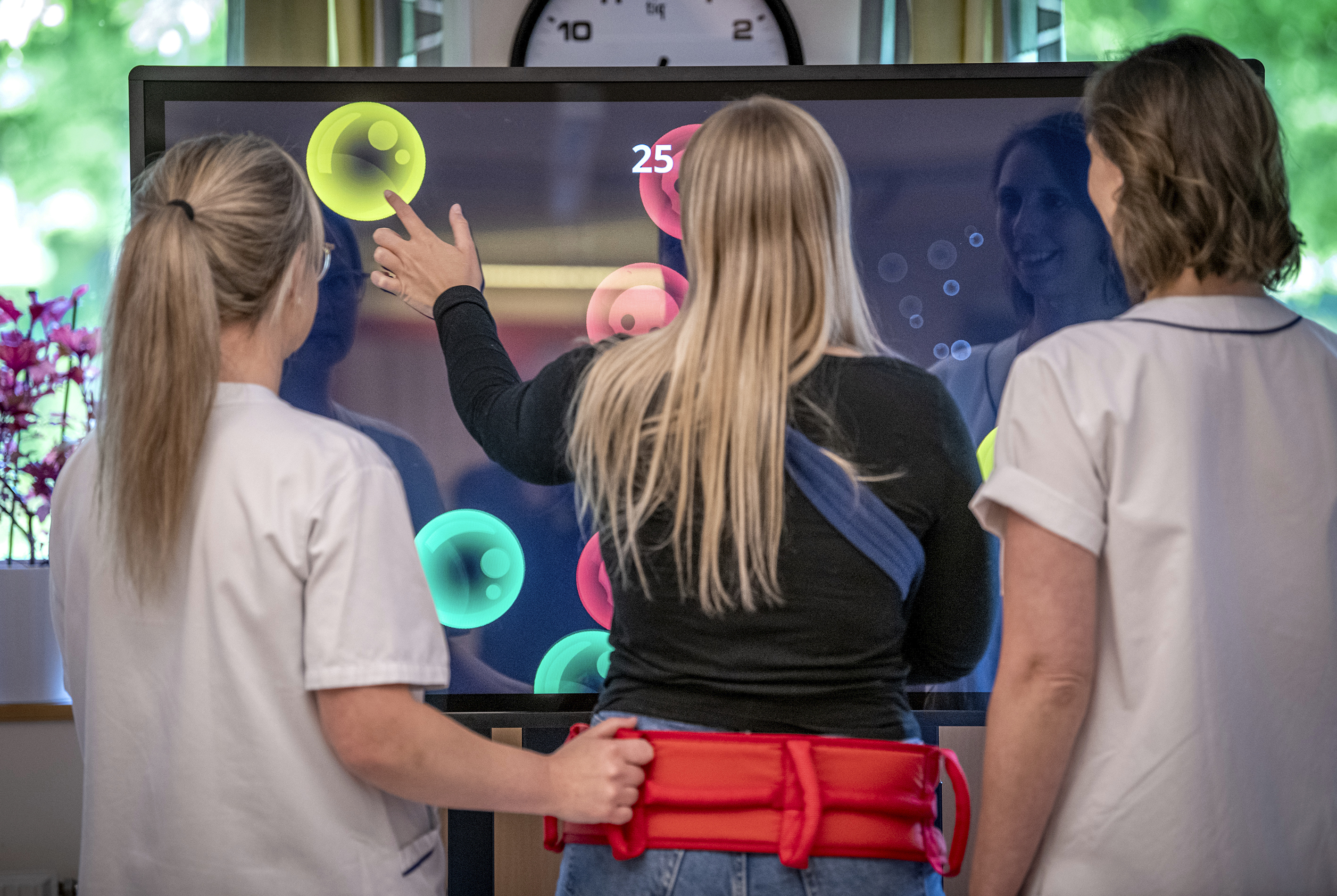 Tre kvinnor med ryggen emot kameran som står fram för en stor skärm där ett spel är öppet och tjejen i motten rör med ett finger på skärmen och spelar spelet