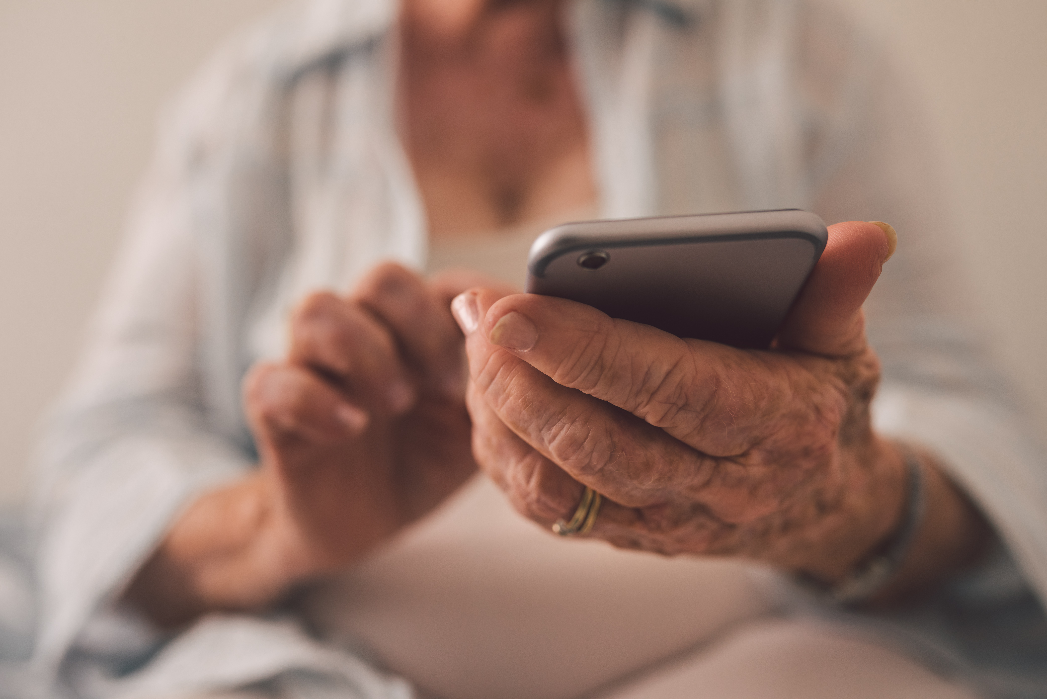 En äldre kvinnas rynkiga hand med två guldringar som håller i en mobiltelefon. Närbild på mobilen och handen.