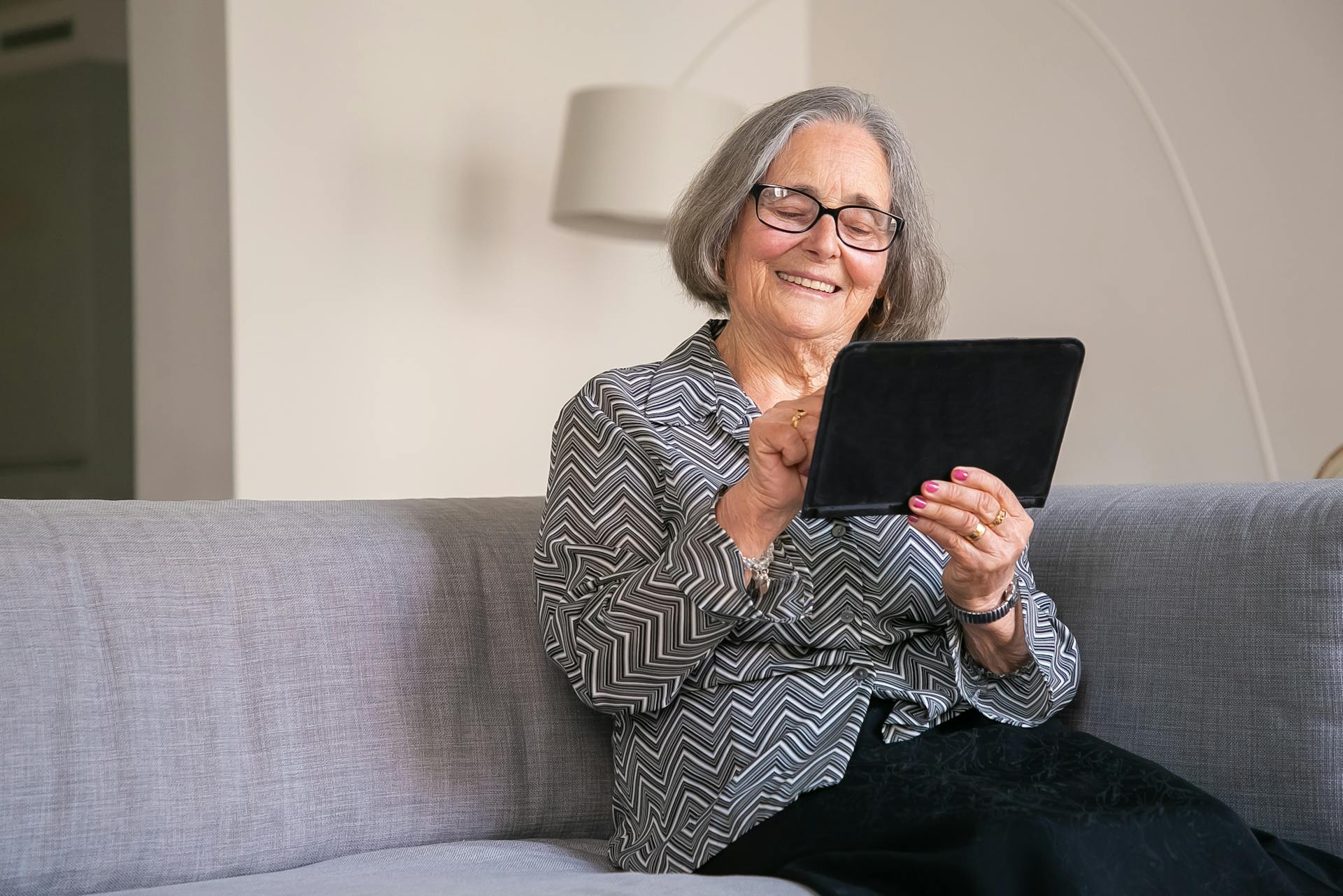 Äldre gråhårig dam med page som sitter och ler framför en skärm som hon håller i handen
