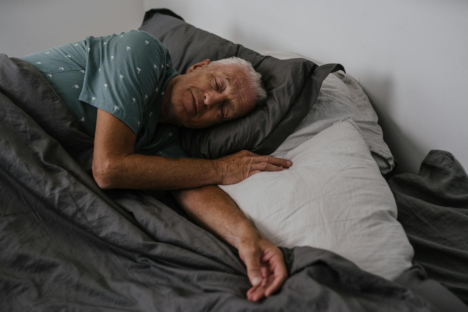 En äldre man som sover i sin säng han har en turkosgrön tröja på sig och grå lakan